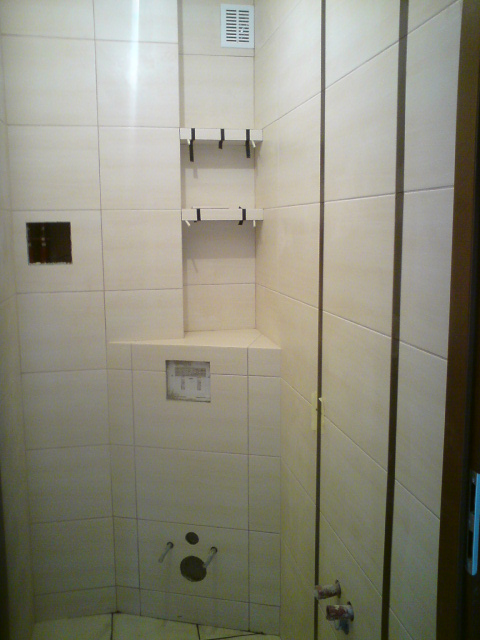 Wykończona łazienka z beżowymi płytkami, wnęką z półkami pod kratką wentylacyjną, zabudowaną spłuczką i wyprowadzonymi przyłączami wodno-kanalizacyjnymi.